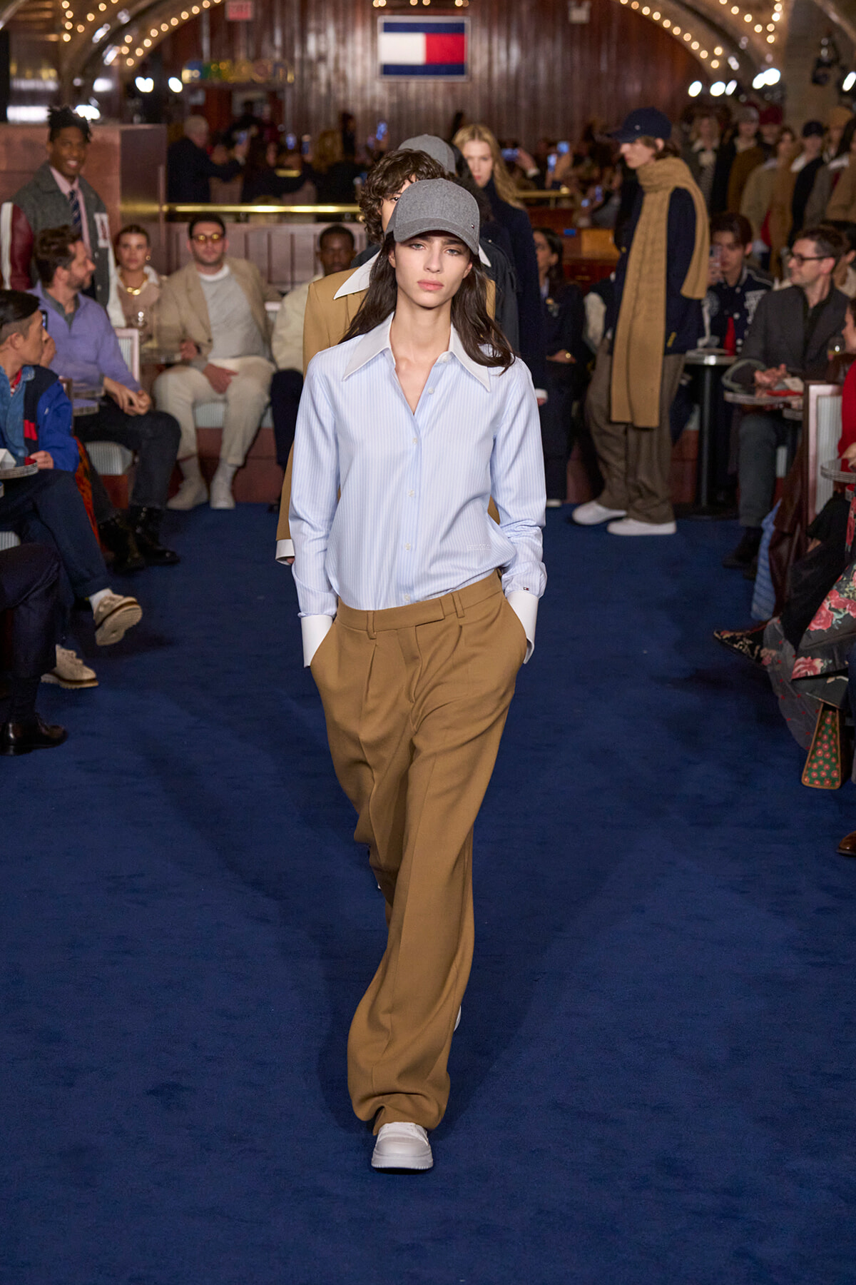 Model walking a fashion show wearing a light blue button-down shirt, tan wide-leg trousers, gray cap, and white sneakers on a blue runway.
