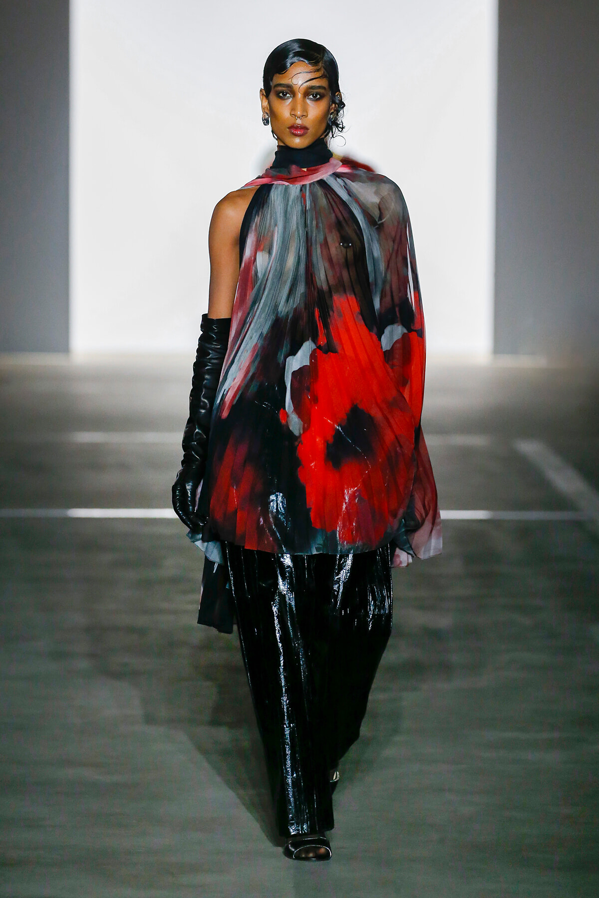 Model walking a fashion runway in a sheer, abstract-red and black cape over a black outfit with long black gloves.