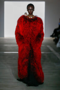 Model on runway wearing a voluminous red fur coat with a gold necklace.