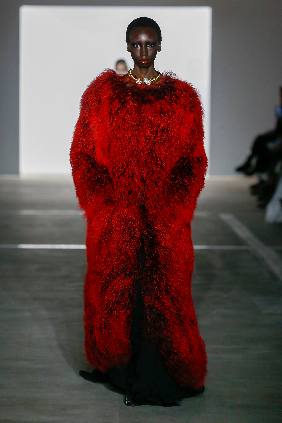 Model on runway wearing a voluminous red fur coat with a gold necklace.