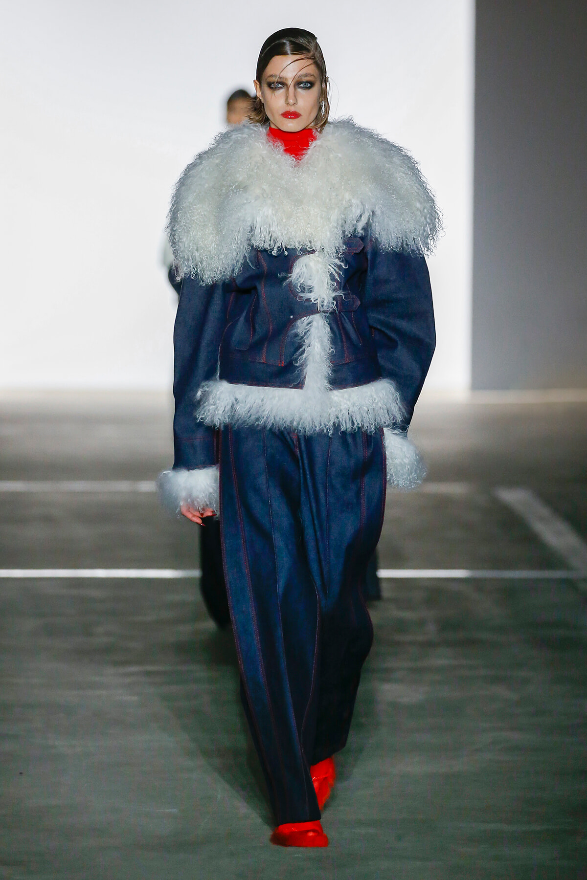 Model walking a fashion runway in a navy coat with oversized white shearling collar and cuffs, red turtleneck, red shoes, and bold makeup.
