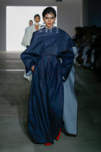 Model walking the runway in a navy oversized cape with red stitching, paired with red pointed shoes and bold earrings.