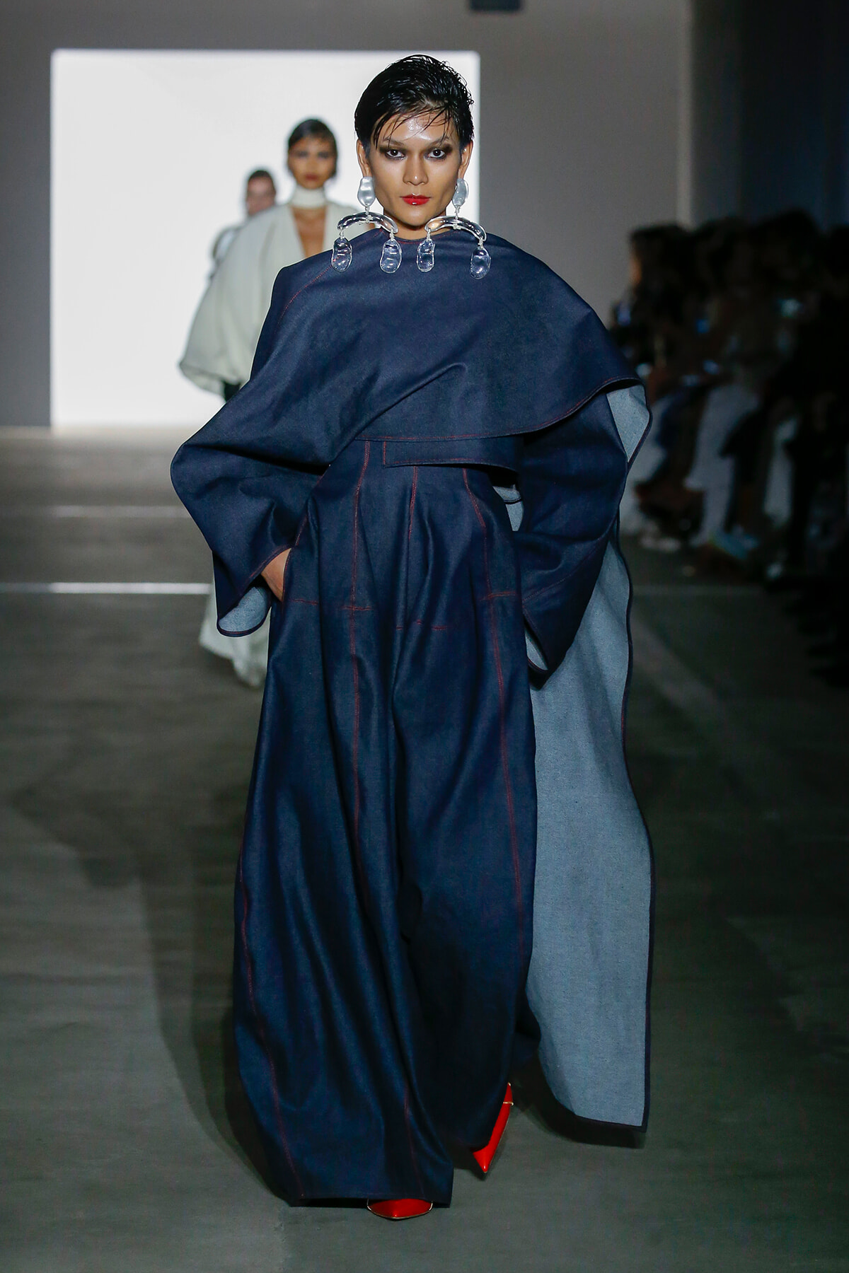 Model walking the runway in a navy oversized cape with red stitching, paired with red pointed shoes and bold earrings.