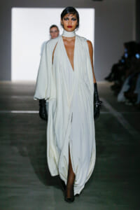 Model walking a runway in a flowing white draped gown with a high neck and cape-like sleeves, paired with black gloves.