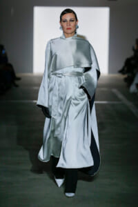 Runway model in a silvery satin cape coat with a tied waist belt, black gloves, and pointed white shoes.