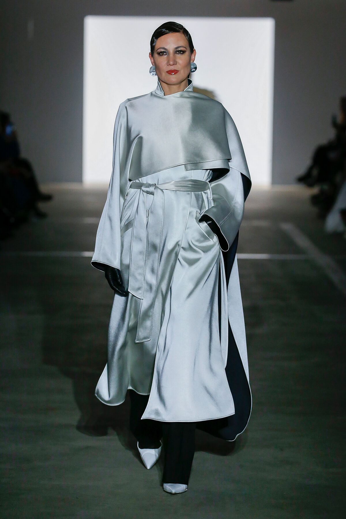 Runway model in a silvery satin cape coat with a tied waist belt, black gloves, and pointed white shoes.