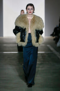 Model walking runway in a oversized beige shaggy fur coat with black leather sleeves, teal satin trousers, and glossy black shoes.