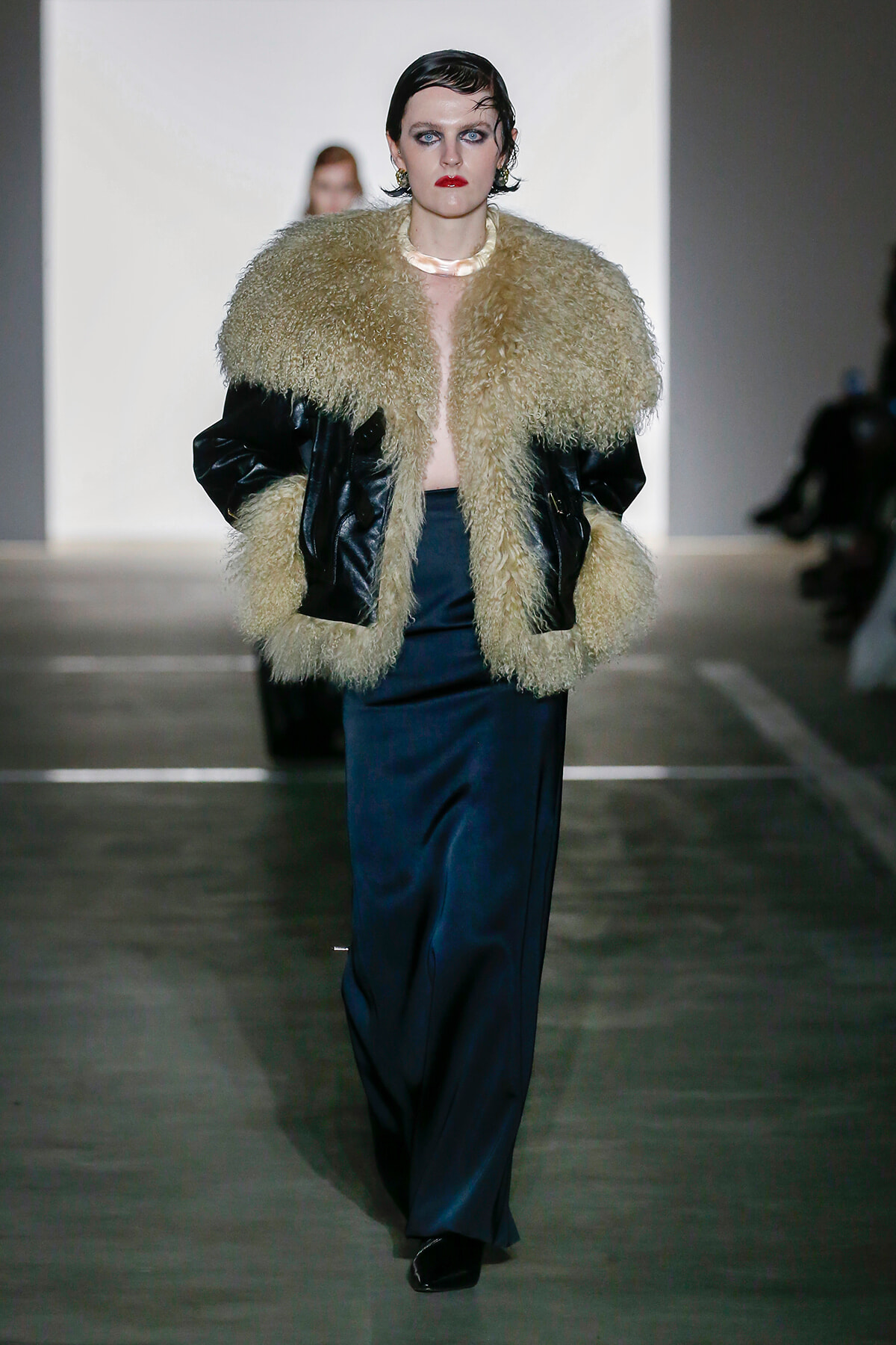 Model walking runway in a oversized beige shaggy fur coat with black leather sleeves, teal satin trousers, and glossy black shoes.