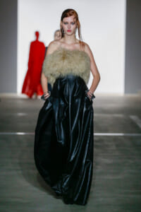 Model walks a runway wearing a black shiny leather gown with a beige fur bustier on a minimalist stage.