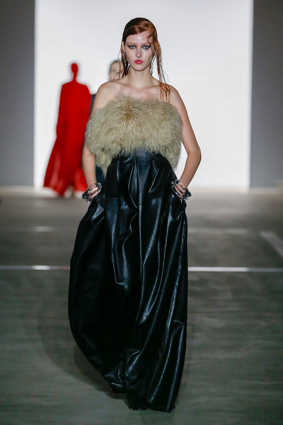 Model walks a runway wearing a black shiny leather gown with a beige fur bustier on a minimalist stage.