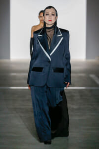 Model walks the runway in a dark navy oversized blazer with white piping and a sheer black turtleneck, paired with matching trousers.
