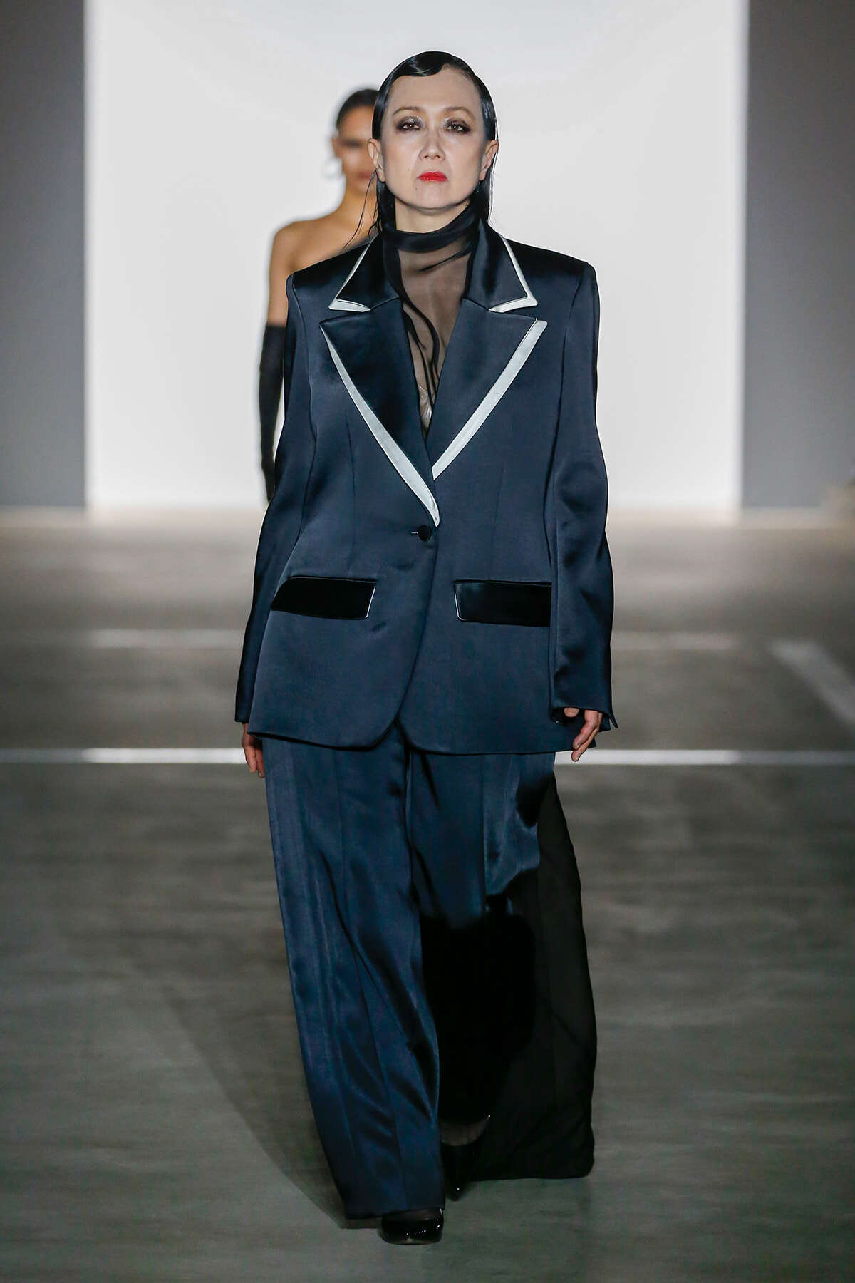 Model walks the runway in a dark navy oversized blazer with white piping and a sheer black turtleneck, paired with matching trousers.