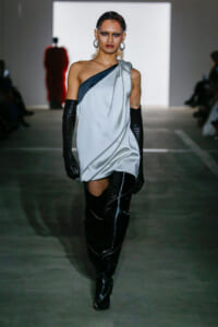 Model walks the runway in a pale gray one-shoulder draped dress with a dark blue accent, paired with long black gloves and thigh-high boots.