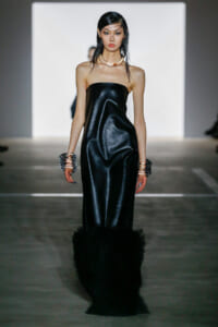 Model walks a runway in a strapless black satin gown with feathered hem, metallic bangles on both wrists, and sleek wet-look hair.