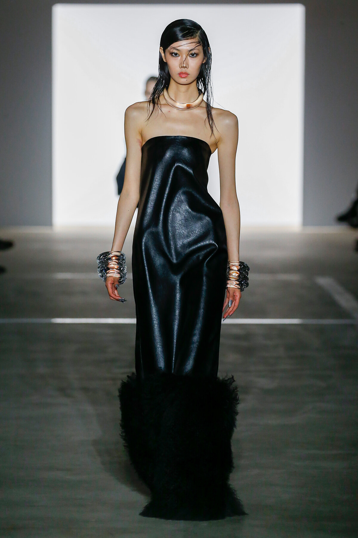 Model walks a runway in a strapless black satin gown with feathered hem, metallic bangles on both wrists, and sleek wet-look hair.