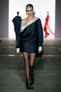 Model struts down a runway in a navy satin one-shoulder mini dress with silver sash and cuffs, bold makeup and slick hair.