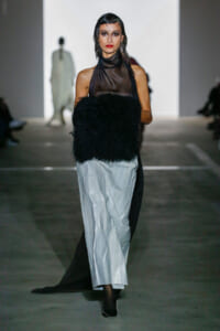 Model walking a runway in a sheer black halter top and light gray pleated pants, carrying a large black fur bag.