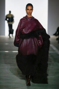 Model walking a runway in a burgundy leather top with fur trim and a black choker, bold makeup.
