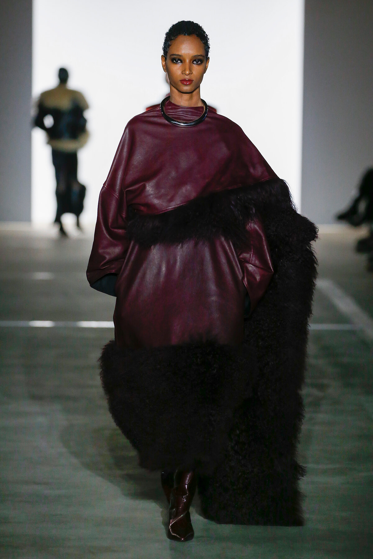 Model walking a runway in a burgundy leather top with fur trim and a black choker, bold makeup.