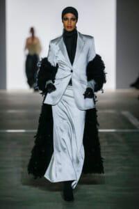 Model walks the runway in a silver satin blazer and matching pants with black feathered cuffs and a black turtleneck beneath.