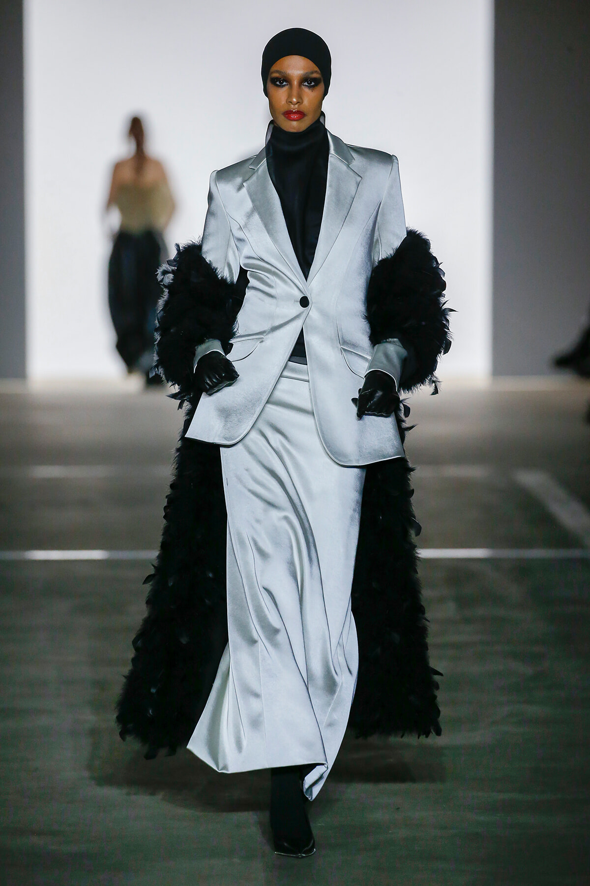 Model walks the runway in a silver satin blazer and matching pants with black feathered cuffs and a black turtleneck beneath.