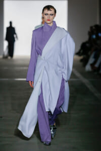 Model walks a fashion runway wearing a lavender and pale blue oversized wrap coat, with slicked-back hair and bold makeup.
