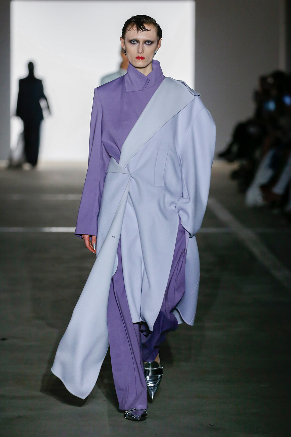 Model walks a fashion runway wearing a lavender and pale blue oversized wrap coat, with slicked-back hair and bold makeup.