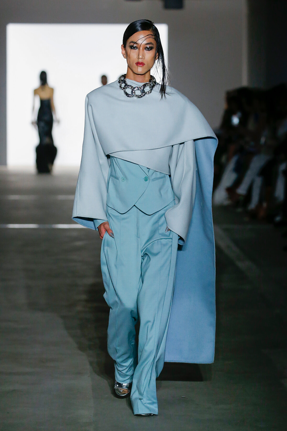 Model walking a runway in a pale blue layered cape and matching suit with a chunky silver chain necklace.