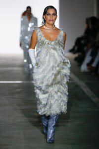 Model walking a runway in a silver feathered gown with long white gloves and blue quilted boots.