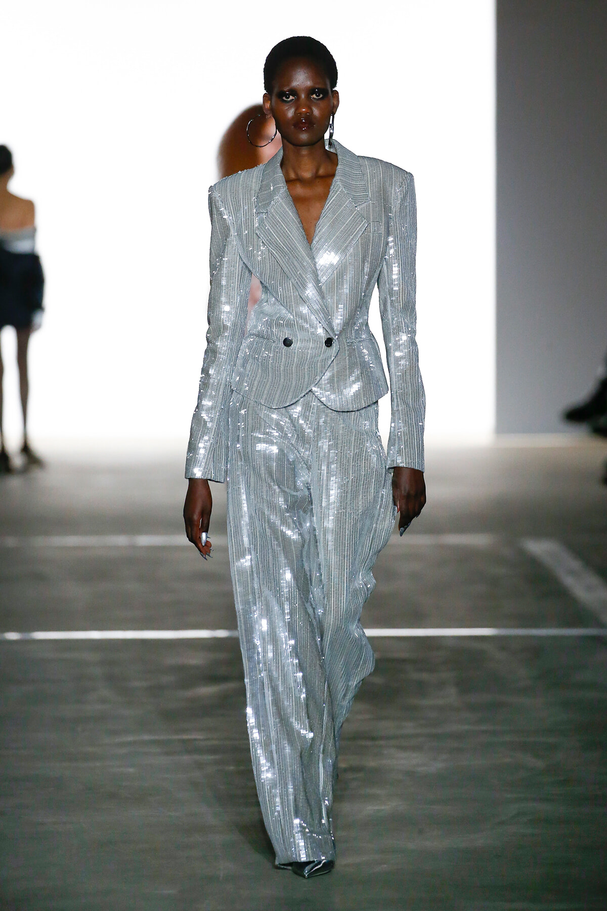 Model walking runway in a metallic silver pinstripe suit with a double-breasted blazer and wide-leg trousers, sequined finish catching the light