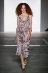 Model walking a runway in a sleeveless, pink-feathered dress with metallic heels and a pale choker, neutral backdrop behind.
