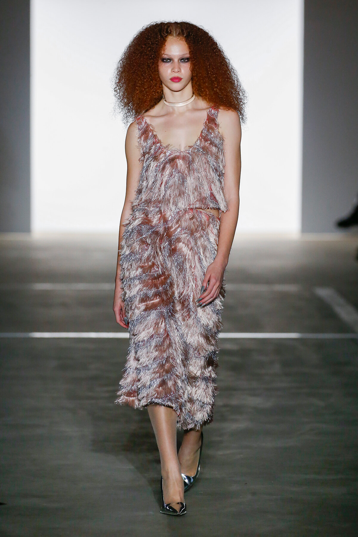 Model walking a runway in a sleeveless, pink-feathered dress with metallic heels and a pale choker, neutral backdrop behind.