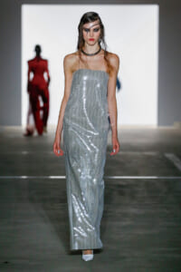 Model walking a runway in a silver strapless sequin gown with white shoes and bold makeup.