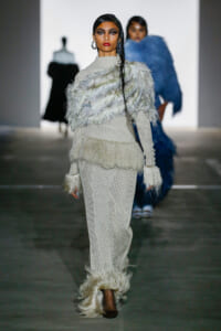 Model walks down a runway in a white knit outfit with layered feather fringe and cuffed sleeves; dark makeup and red lips, bold earrings visible.