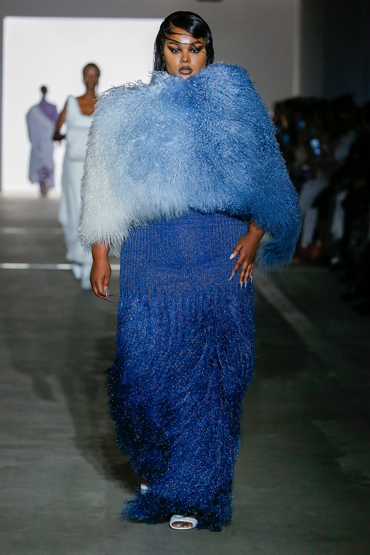 Model on a runway wearing a shimmering, beaded blue gown with a large pale-blue fluffy cape.