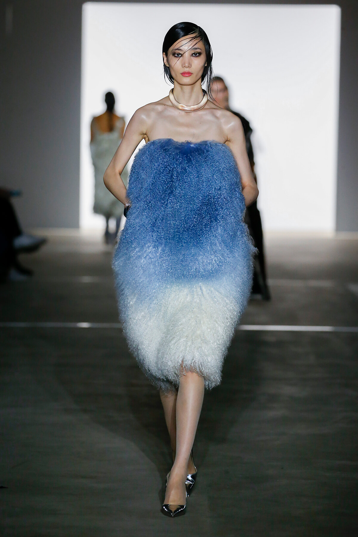 Model walking a runway in a strapless, fluffy dress that fades from blue to white, with a light pink necklace and silver heels.
