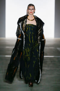 Model on a runway wearing a dark, patterned dress and a fur coat, accessorized with a gold choker and earrings.