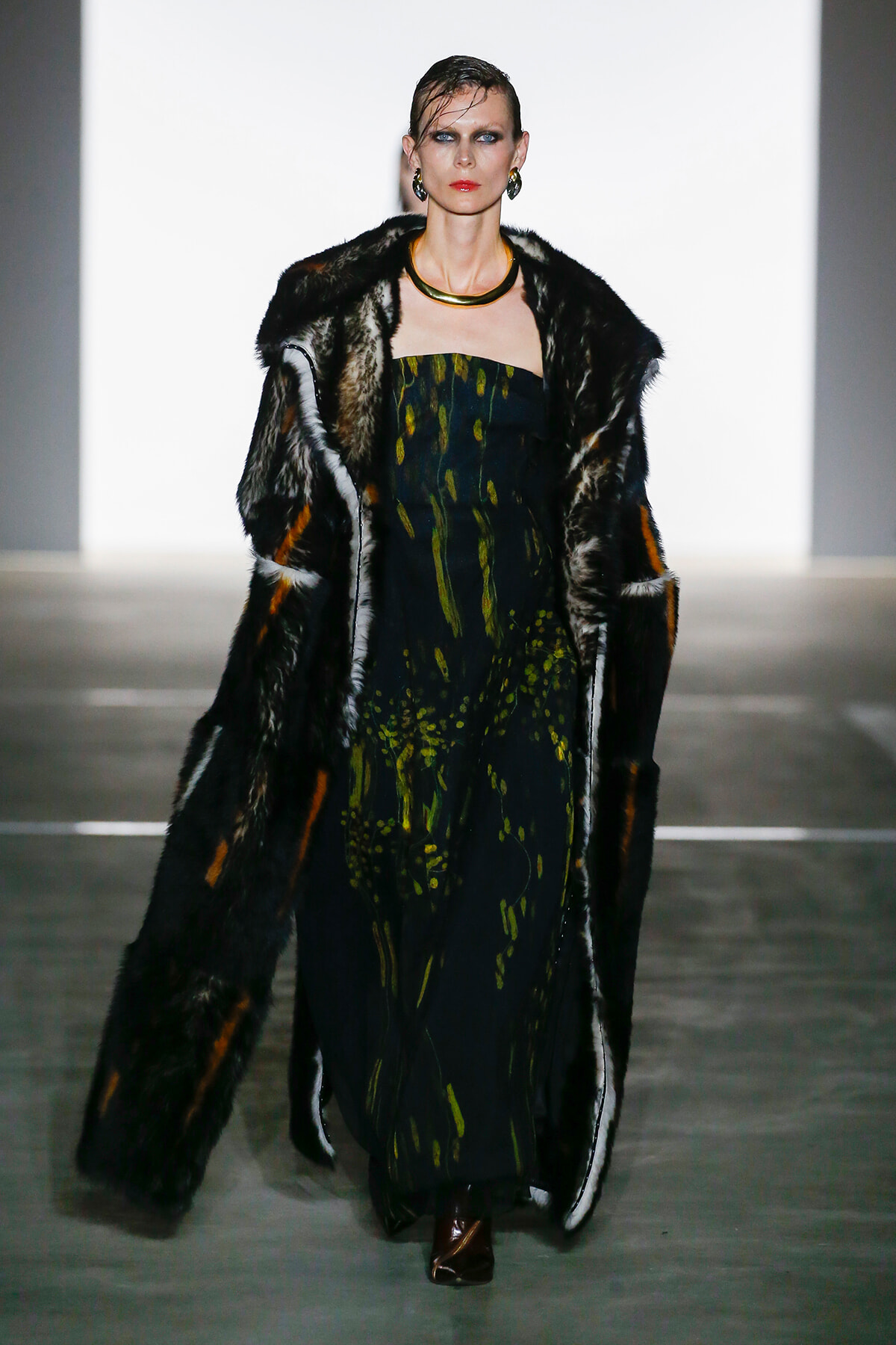 Model on a runway wearing a dark, patterned dress and a fur coat, accessorized with a gold choker and earrings.