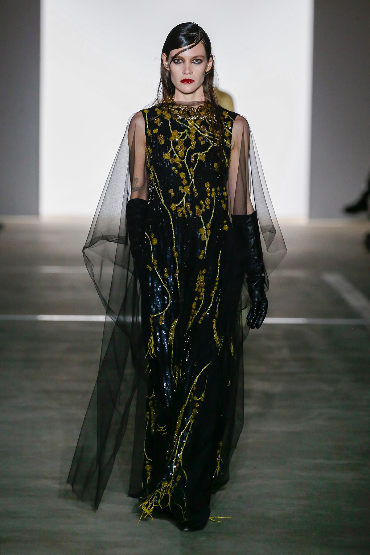 Model walking runway in a floor-length black gown with gold embroidery and sheer cape sleeves, wearing black gloves.