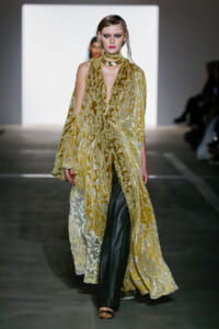 Runway model in a gold patterned sheer cape over dark pants, high makeup, walking the runway in a fashion show.