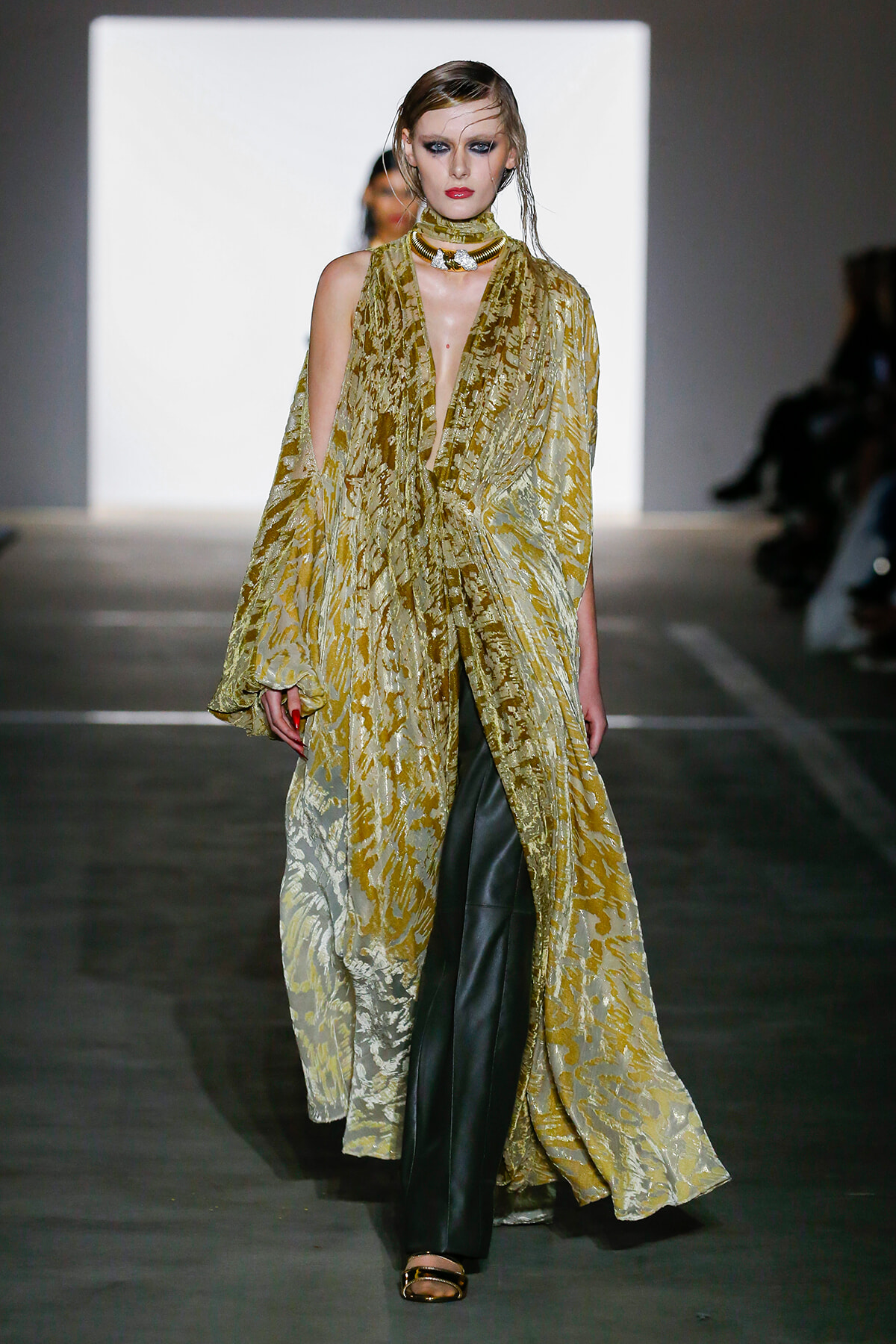 Runway model in a gold patterned sheer cape over dark pants, high makeup, walking the runway in a fashion show.