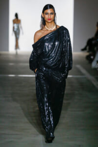 Model walking a fashion runway in a one-shoulder, black sequined jumpsuit with a gold choker and wet hairstyle, spotlighted on the catwalk.