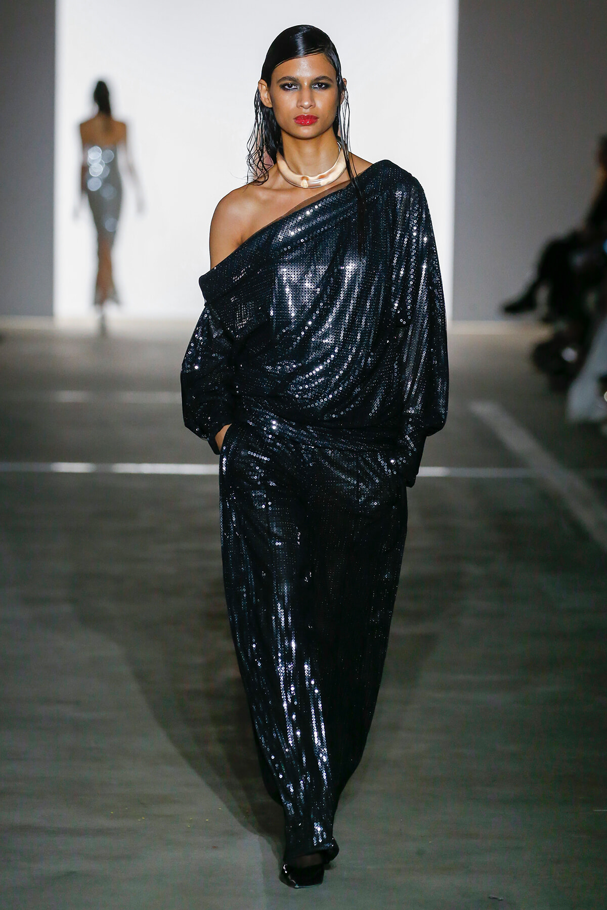 Model walking a fashion runway in a one-shoulder, black sequined jumpsuit with a gold choker and wet hairstyle, spotlighted on the catwalk.