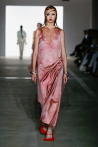 Runway model in a pink satin draped gown with a deep V neck, silver bracelets, and red fuzzy heeled sandals.