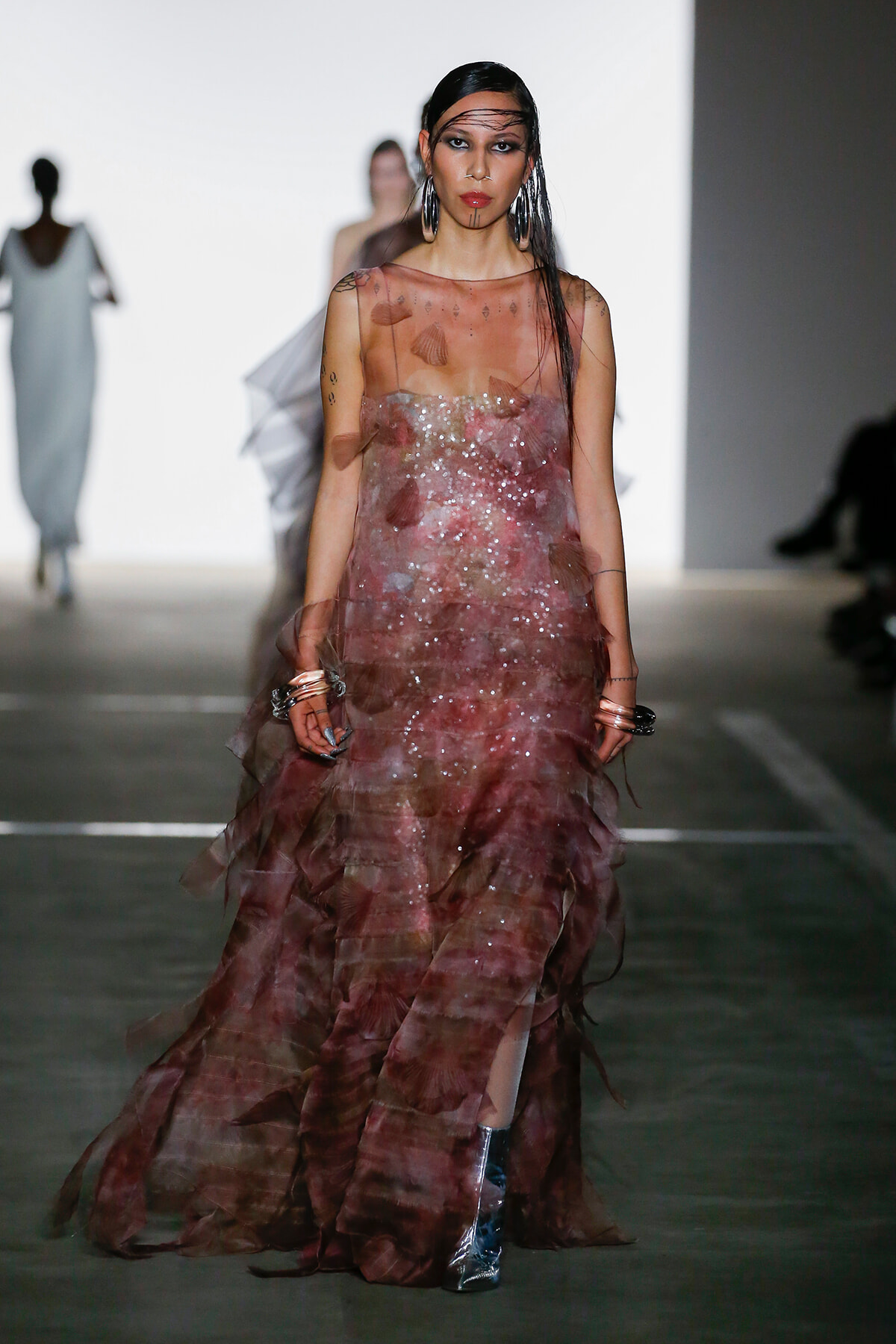 Model walking a fashion runway in a sheer pink-brown sequined gown with a layered, flowing skirt and metallic boots; bold makeup and long dark hair.