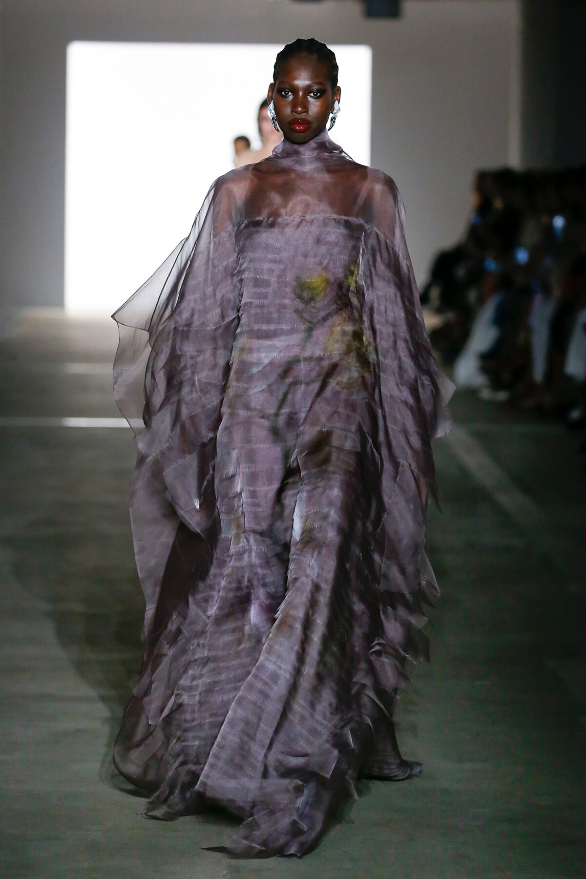 Model walks the runway in a flowing lavender sheer gown with layered ruffles and a floral print, on a dimly lit catwalk.