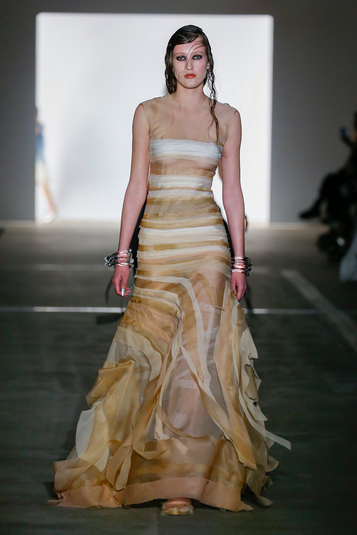 Model walks the runway in a sheer, striped beige gown with a fitted bodice and voluminous tiered skirt, accessorized with chunky bracelets on both wrists.