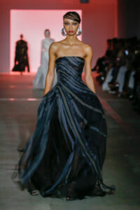 Model walks a runway in a strapless, dark striped gown with a voluminous, layered skirt; pink stage backdrop and audience seated along the runway.
