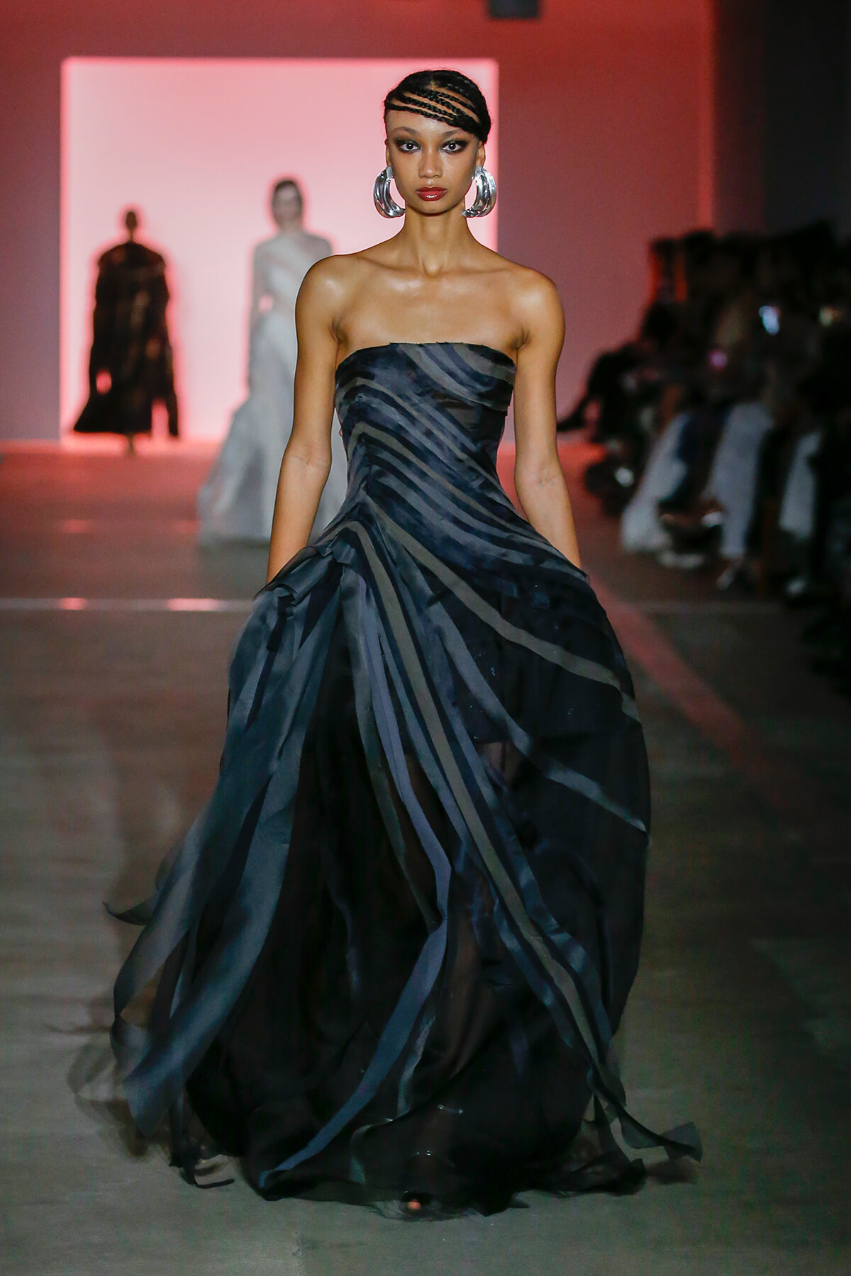 Model walks a runway in a strapless, dark striped gown with a voluminous, layered skirt; pink stage backdrop and audience seated along the runway.