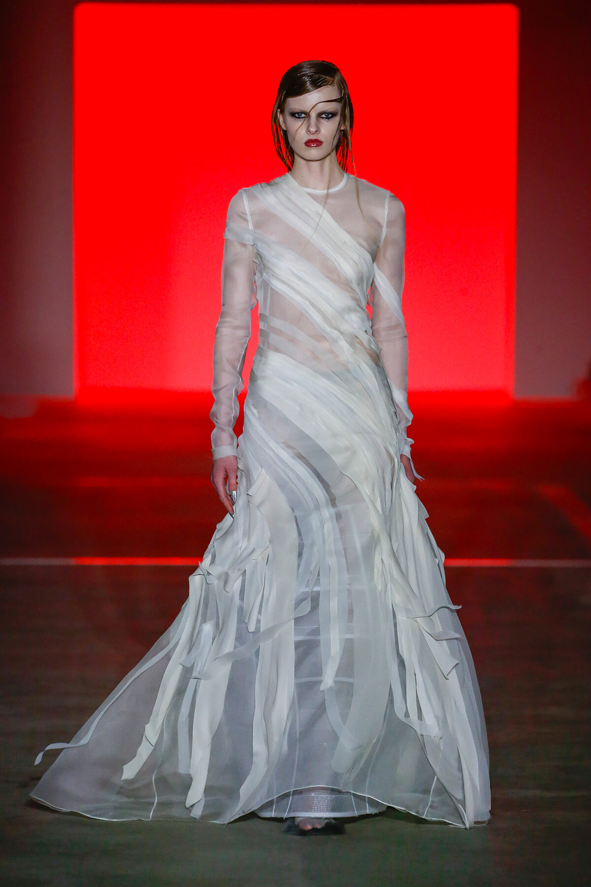 Model walking the runway in a layered, sheer white gown with ribbon-like strips against a bright red backdrop.